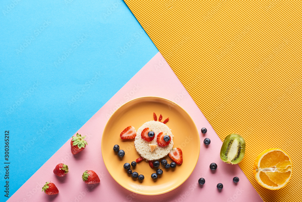 Fototapeta top view of plate with fancy animal made of food for childrens breakfast on blue, yellow and pink background