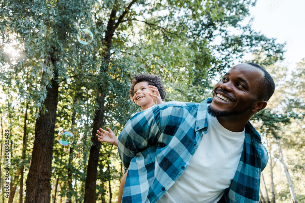 low angle view of happy african american piggybacking cute curly son near soap bubbles in park low angle view of happy african american piggybacking cute curly son near soap bubbles in park