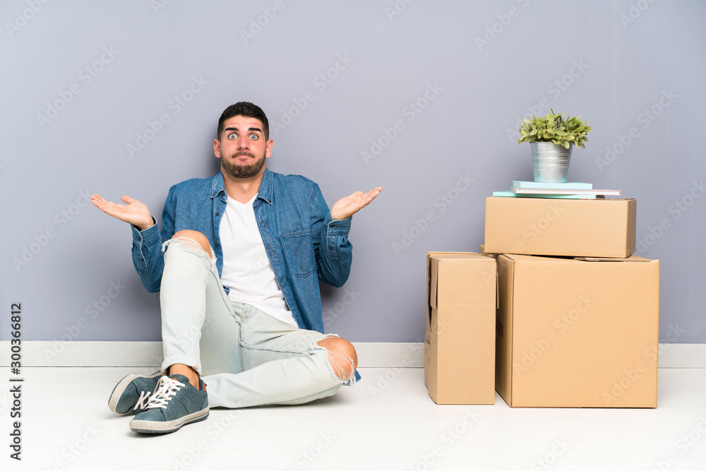 Handsome young man moving in new home among boxes having doubts with confuse face expression Handsome young man moving in new home among boxes having doubts with confuse face expression
