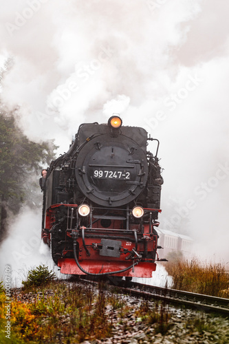 Antique and original Harz steam locomotive passing through the fog and steam during a moody autumn day with orange trees and dark smoke (Harz, Germany)