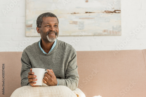 senior, positive african american man sitting with blanket on knees and holding cup of warming drink