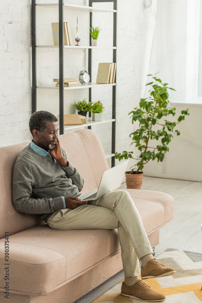 thoughtful african american man sitting on sofa in living room and using laptop thoughtful african american man sitting on sofa in living room and using laptop