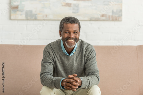 senior african american man sitting on sofa with clenched hands and smiling at camera
