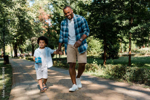 happy african american father holding hands with son while walking on path in park