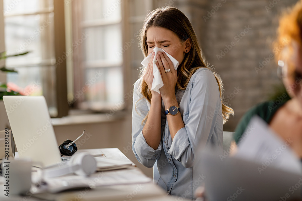 Young businesswoman using a tissue while sneezing in the office. - obrazy, fototapety, plakaty 