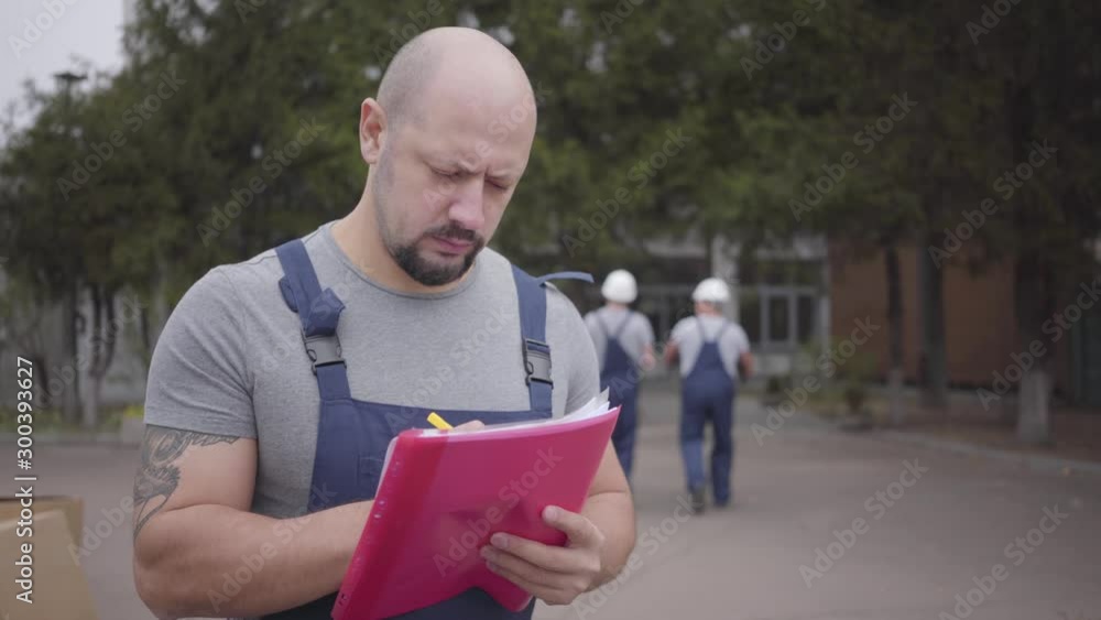 Portrait of an adult bearded Caucasian man writing in the waybill as two workers walking away with packages. Working in the moving service. Portrait of an adult bearded Caucasian man writing in the waybill as two workers walking away with packages. Working in the moving service.