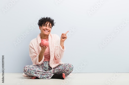 Young african american woman sitting with a piggy bank smiling cheerfully pointing with forefinger away. © Asier