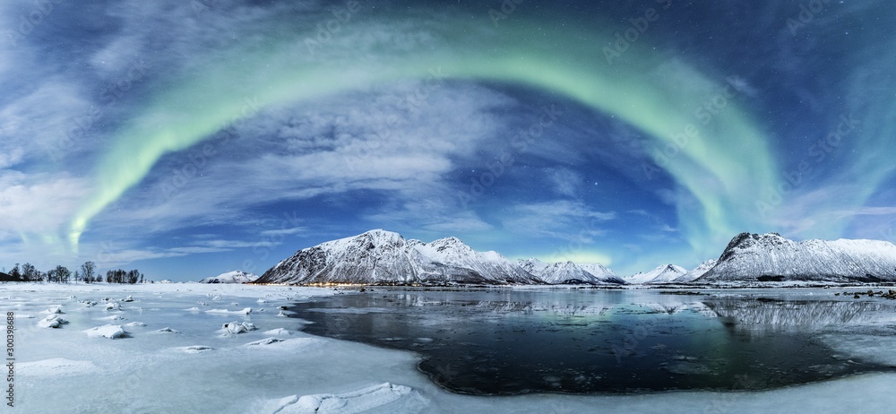 Wide shot of the arch shaped northern lights over a frozen sea with the snow covered mountains - obrazy, fototapety, plakaty 