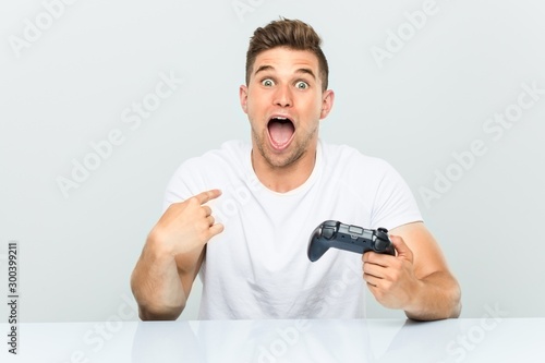 Young man holding a game controller surprised pointing at himself, smiling broadly.
