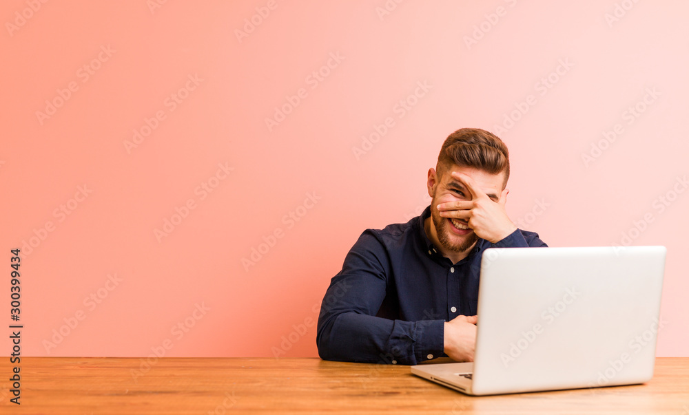 Young man working with his laptop blink at the camera through fingers, embarrassed covering face. Young man working with his laptop blink at the camera through fingers, embarrassed covering face.