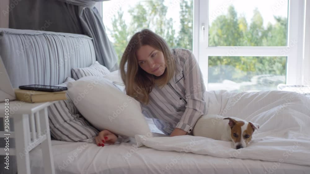 Tracking of beautiful woman with cute dog waking up in morning and turning off alarm clock on mobile phone, then stretching in bed. Blue jiu-jitsu gi with black belt hanging on chair Tracking of beautiful woman with cute dog waking up in morning and turning off alarm clock on mobile phone, then stretching in bed. Blue jiu-jitsu gi with black belt hanging on chair