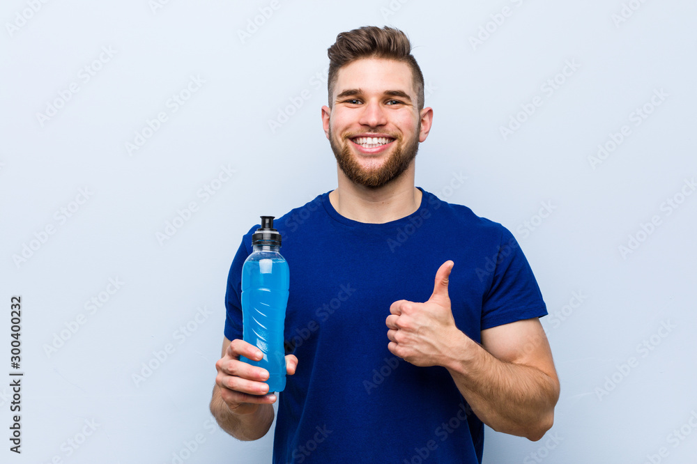 Young caucasian sportsman holding an isotonic drink smiling and raising thumb up - obrazy, fototapety, plakaty 
