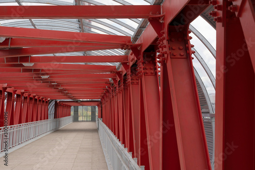 Pedestrian crossing, construction of red metal structures. The roof is made of steel channels connected to each other. Red iron beams on boltsю