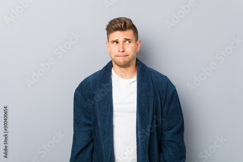 Young caucasian man wearing pajama confused, feels doubtful and unsure.