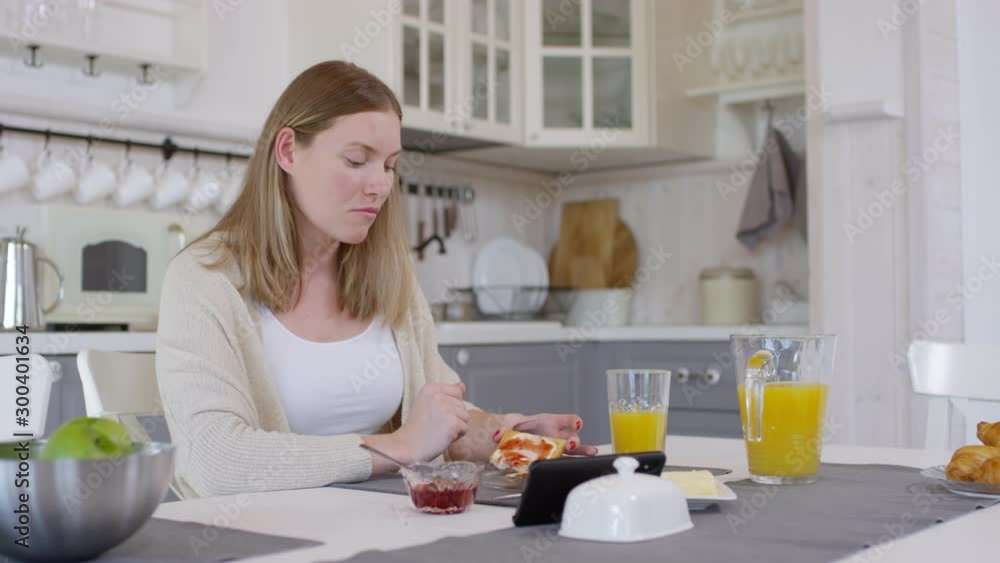 Tracking of cheerful woman sitting at table and watching video on mobile phone while eating toast with jam and butter and drinking orange juice in morning Tracking of cheerful woman sitting at table and watching video on mobile phone while eating toast with jam and butter and drinking orange juice in morning