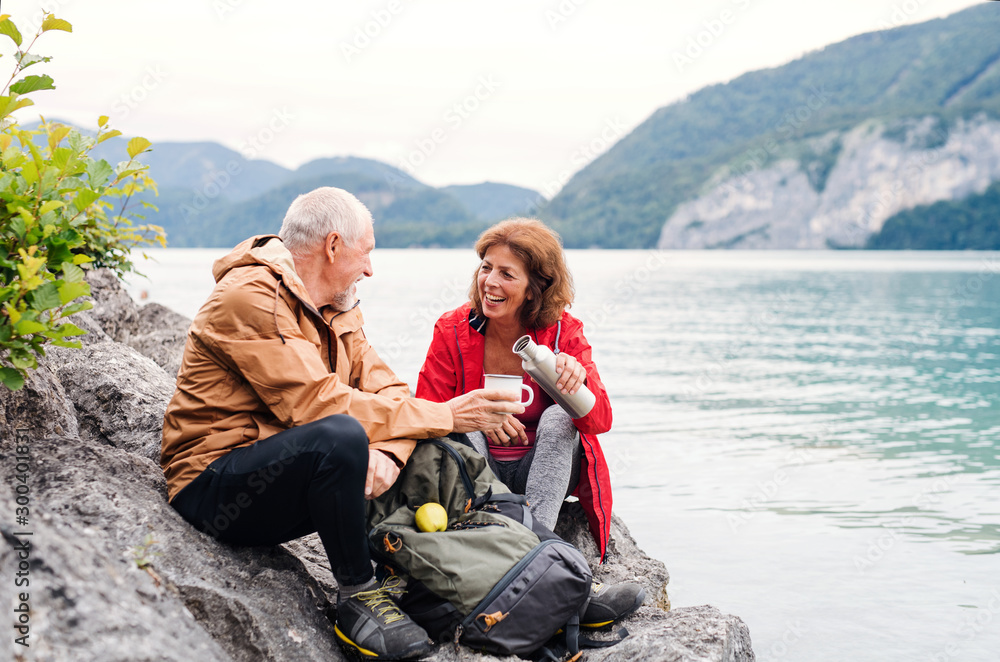 A senior pensioner couple hiking by lake in nature, sitting and resting. - obrazy, fototapety, plakaty 