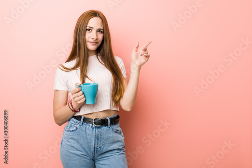 Young caucasian woman holding a cup smiling cheerfully pointing with forefinger away.