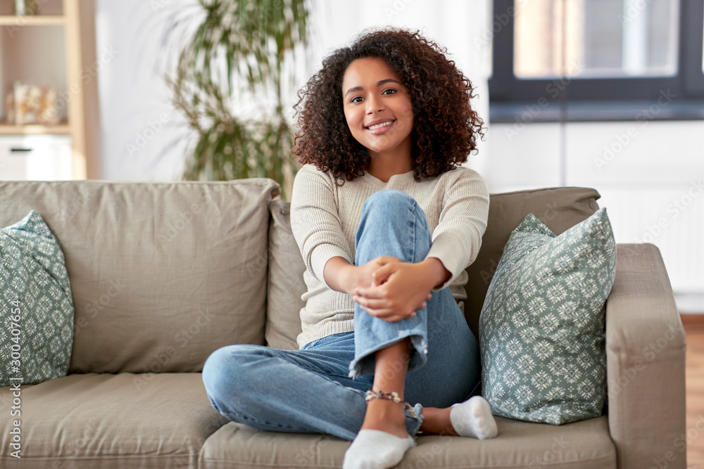 people, race, ethnicity and portrait concept - happy african american young woman sitting on sofa at home - obrazy, fototapety, plakaty 