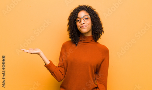 young pretty black woman feeling happy and smiling casually, looking to an object or concept held on the hand on the side against orange wall