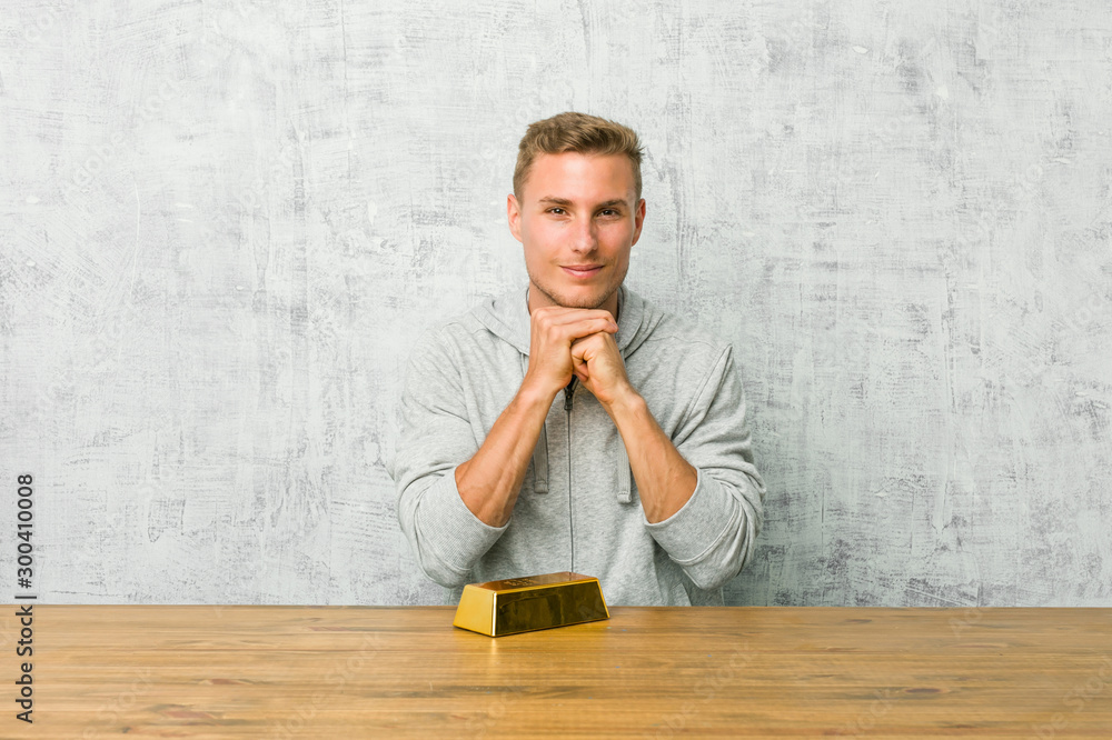 Obraz premium Young handsome man holding a gold ingot on a table keeps hands under chin, is looking happily aside.
