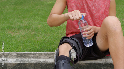 Cropped image of young teenager boy's wears supportive knee brace, relaxing and open fresh water bottle to drink during jogging in public park at morning time, exercising and health care concept 