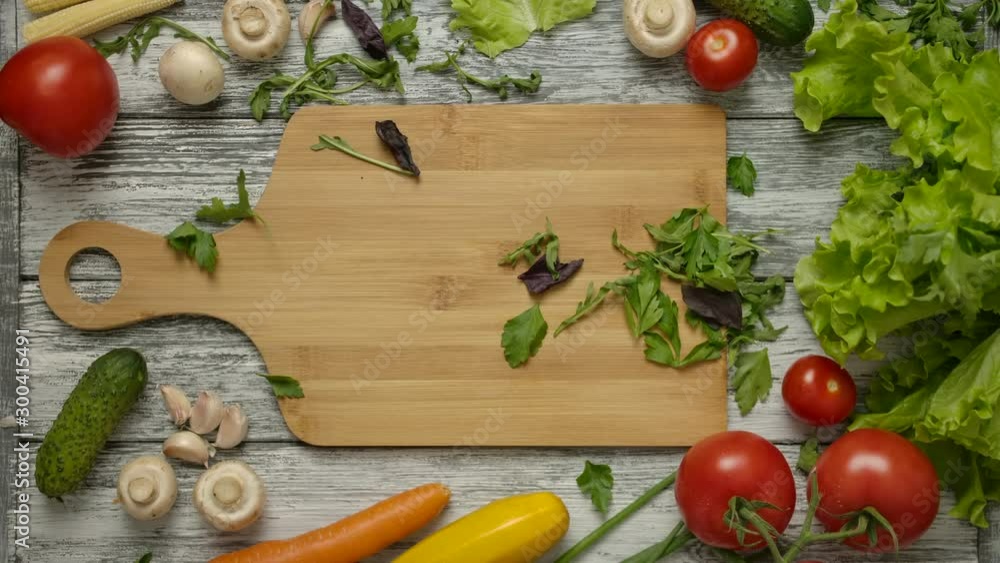 Overhead slow motion shot of parsley and basil leaves falling on cutting board, rack focus. From above shot of wooden kitchen table with vegetables. Overhead slow motion shot of parsley and basil leaves falling on cutting board, rack focus. From above shot of wooden kitchen table with vegetables.