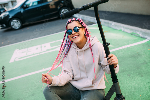 City portrait of handsome hipster girl with colored afro braids riding an electric scooter in the city.
