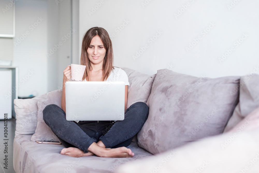 Adult woman sitting on a sofa while using laptop and holding cup of tea Adult woman sitting on a sofa while using laptop and holding cup of tea
