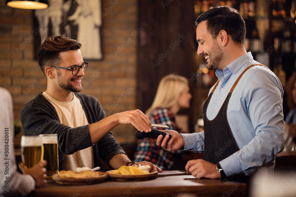 Happy waiter charging a bill via contactless payment in a pub.