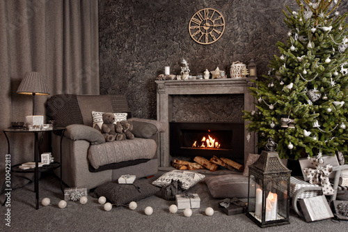 Modern cozy interior in gray shades with an armchair in front of fireplace and decorated Christmas tree. On the chair there are nice knitted blanket, cushions and Teddy bear. Candles, gifts and other 