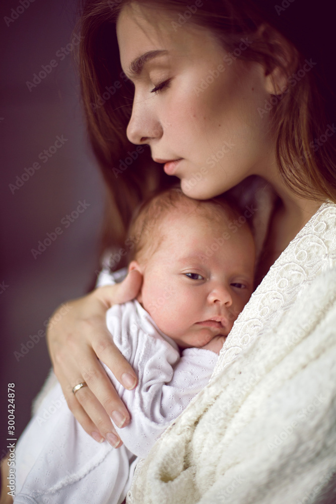 mother in a white robe holds a small newborn daughter against the wall mother in a white robe holds a small newborn daughter against the wall