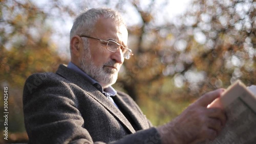 Handsome grandfather with a beautiful beard in a gray jacket sits on a bench in the park and reads a newspaper. Senior gray-haired man in glasses