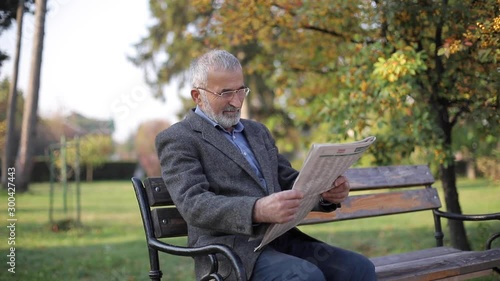 Bearded elderly man in glasses reading newspaper in the autumn park. Handsome gray-haired man sitting on the bench early in the morning