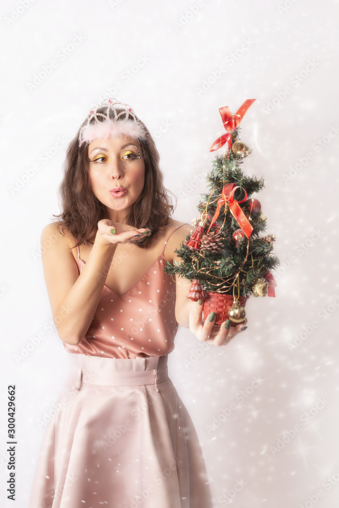 A beautiful young girl in a fairy fairy or Princess costume holds a Christmas tree in her hand and blows on it on a white background. Happy New Year! Empty space for a postcard.