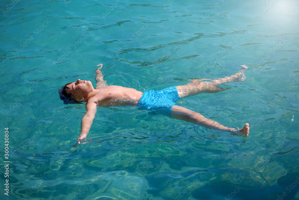 European boy is laying in blue clear sea water in the form of sea star. He is relaxing during his summer holidays.
