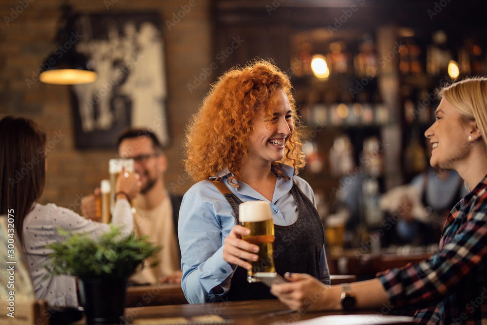 Happy waitress talking to a woman while giving her a glass of beer in a pub. - obrazy, fototapety, plakaty 