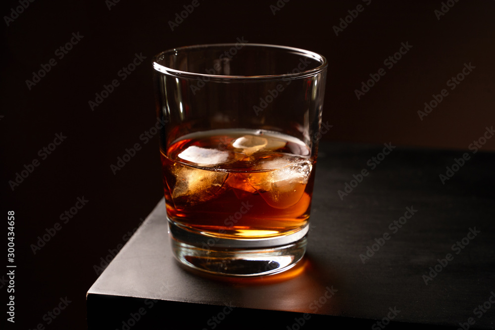 A glass of whiskey with ice on a wooden table. Low key. Horizontal orientation. A glass of whiskey with ice on a wooden table. Low key. Horizontal orientation.