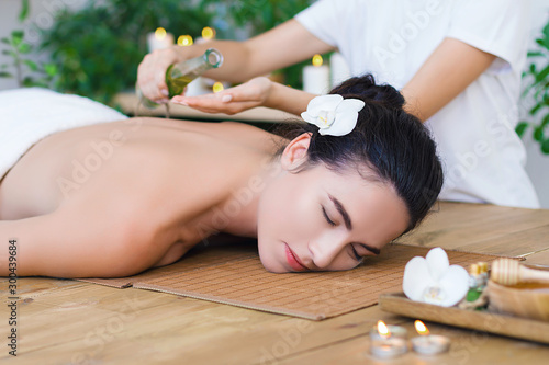 Young pleased woman is getting thai massage, therapy. Female master is pouring massaging oil in palms. Brunette girl is lying on couch in light spa ayurveda salon. Relax and health care concept.