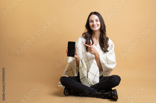 An image of a young beautiful woman in a white sweater and plaid who is pointing at the phone on an isolated orange background