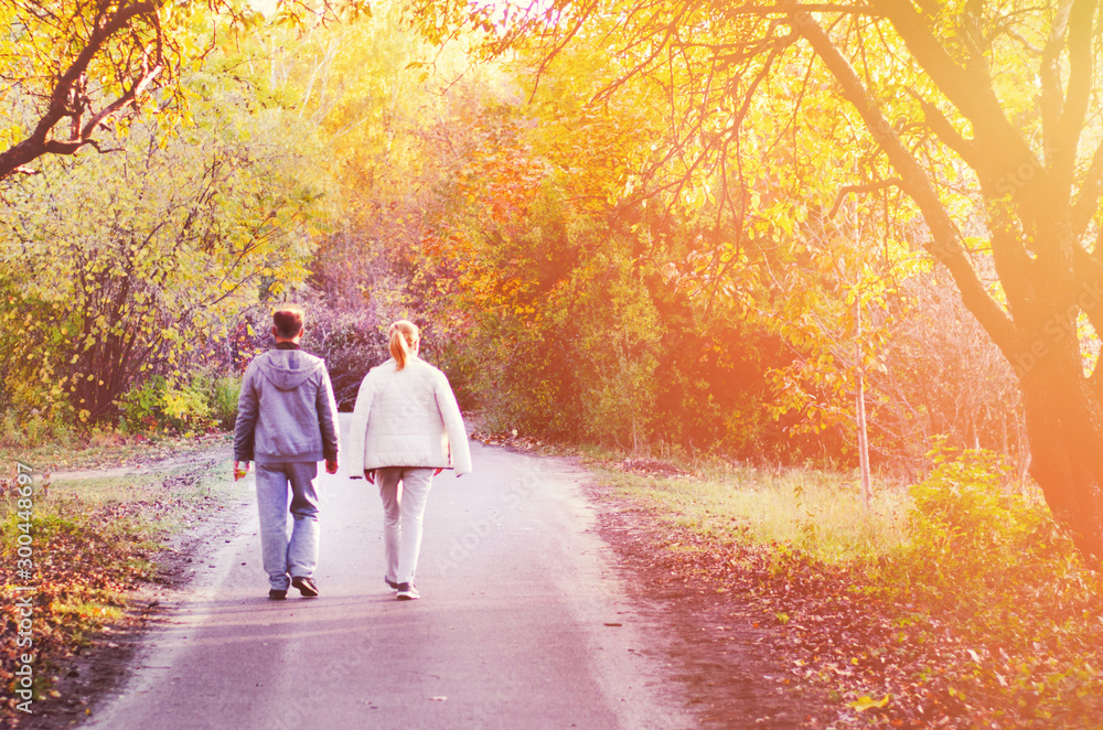Two people, man and woman walking in autumn park with yellow, red and orange trees, falling leaves and bright sun beams. Seasonal concept. Fading and lifestyle concept. Back side view. - obrazy, fototapety, plakaty 