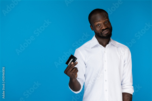 playful looking forward afroamerican man in white shirt is holding credit card in the right hand on the blue background photo