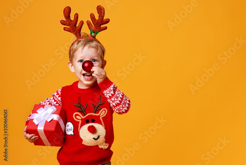 happy funny child boy in red Christmas reindeer costume with gift on yellow   background