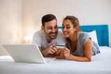 Beautiful young amorous couple using laptop, on bed. Happy young couple shopping online while lying on their bed. Young couple surfing on internet and shopping with laptop on bed.