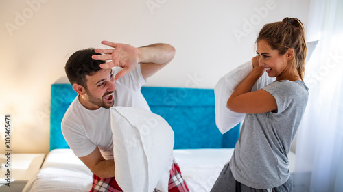 Happy young couple is having fun in bed. Enjoying the company of each other. Pillow battle. Cheerful loving couple is fighting by pillows in bed. They are sitting and laughing