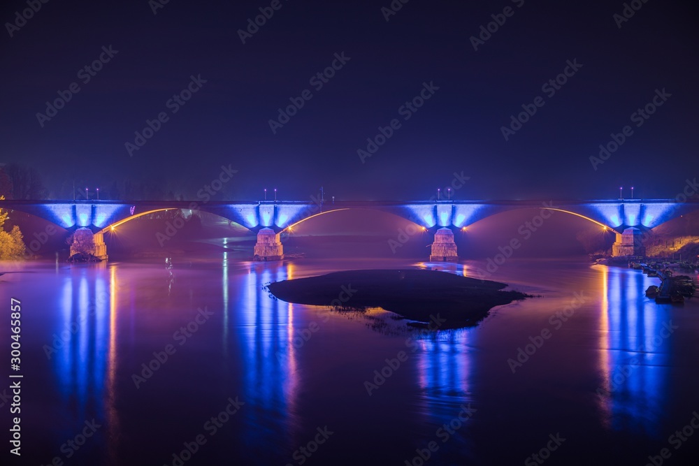 Fototapeta Concrete bridge with beautiful blue lights reflected in the lake at night in Pavia, Italy