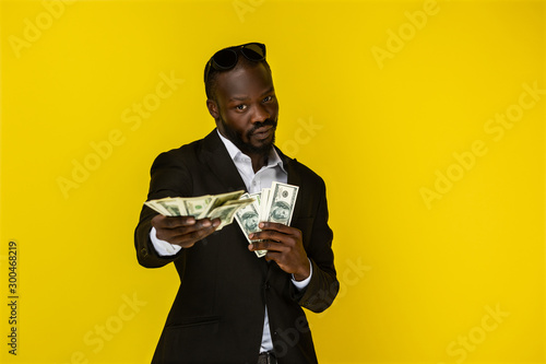 bearded luxury young afroamerican guy is holding lots of money in both hands and look in front of him in sunglasses on the top of the head and black suit on the yellow background