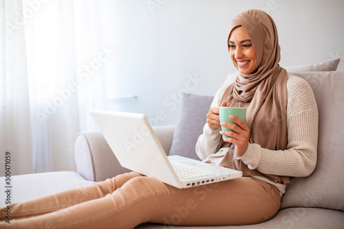 Enjoying time at home. Beautiful young smiling Muslim woman working on laptop and drinking coffee while sitting in a sofa at home. Young Muslim woman using laptop while sitting on comfortable sofa