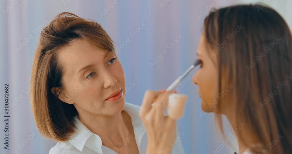 Makeup artist does professional makeup of model in beauty studio. Master applies shadow with a brush on the eyelids of the girl. Portrait view. Makeup artist does professional makeup of model in beauty studio. Master applies shadow with a brush on the eyelids of the girl. Portrait view.