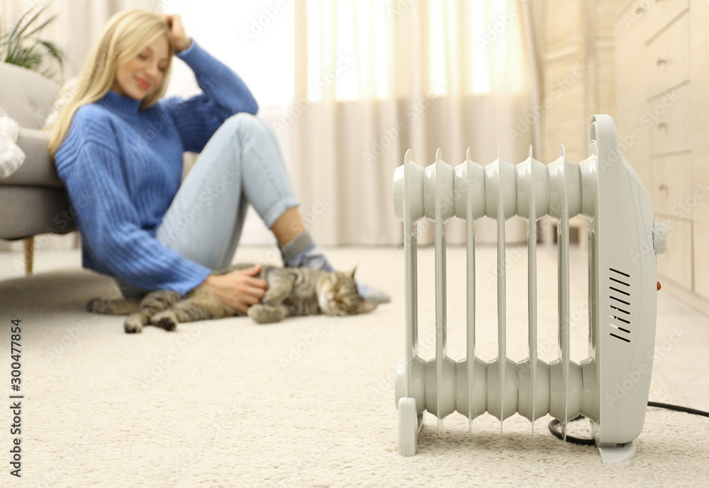 Electric heater and blurred young woman with cute tabby cat on background. Space for text - obrazy, fototapety, plakaty 