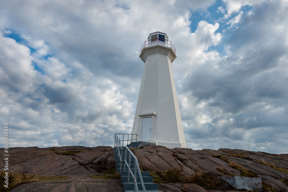 An onshore lighthouse built on a rocky foundation with steps and a handrail leading up to the structure. A stark white concrete tower with a light beacon on top. Blue sky and clouds in the background 